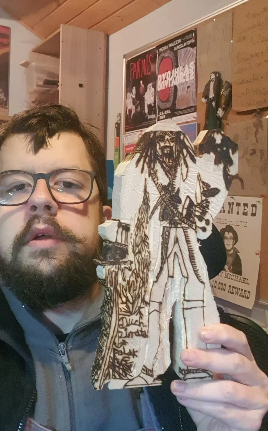Michał w okularach, z brodą, trzyma wypaloną figurkę pirata przed sobą; w tle ściana pracowni