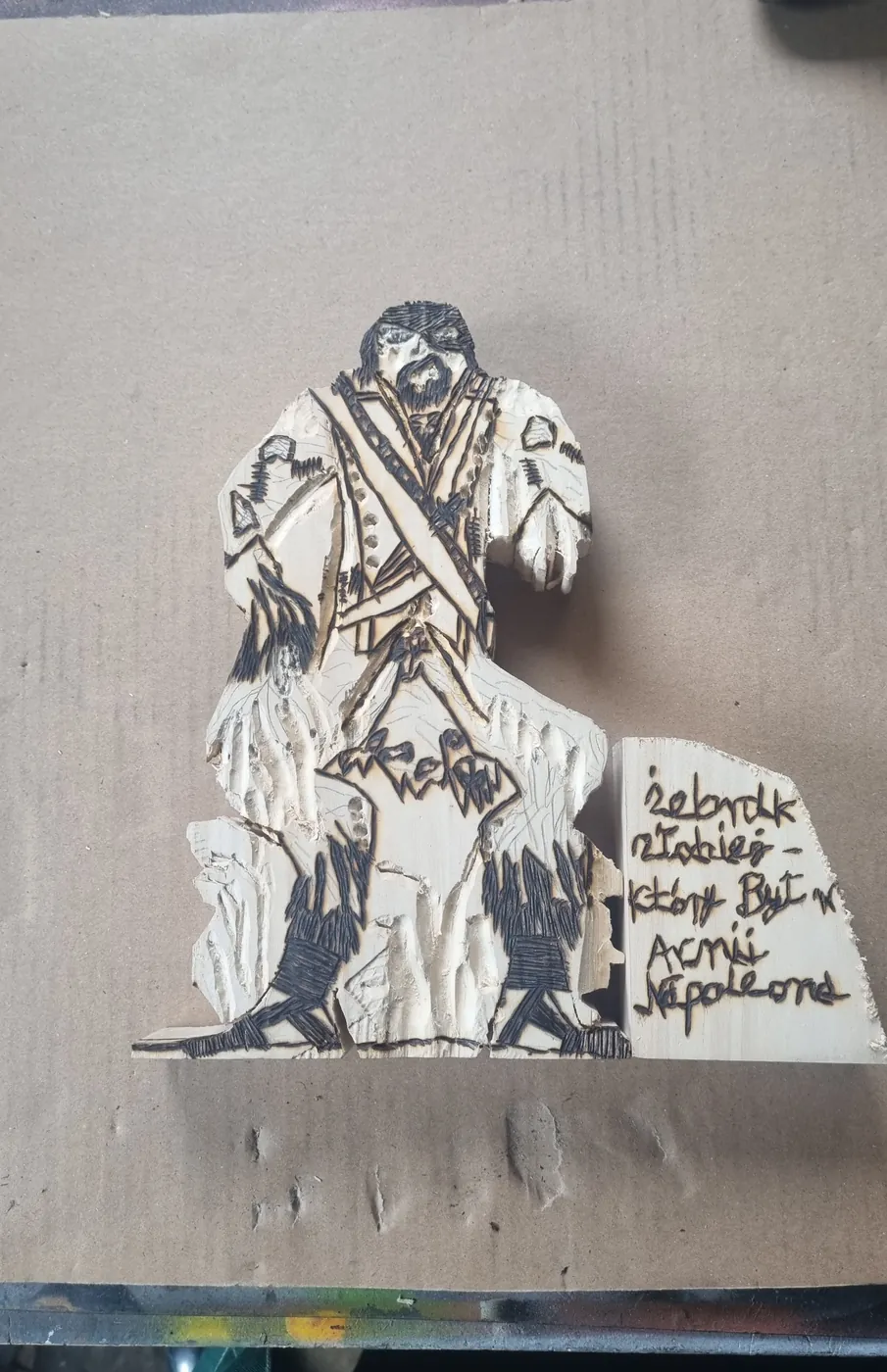 Figurka z wypalonymi ciemnymi konturami; obok tabliczka z wypalonym napisem Żebrak Złodziej, który był w armii Napoleona