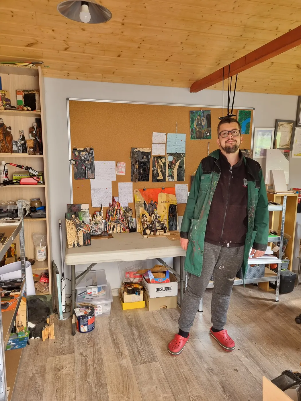 Michał Walczyk stoi w pracowni, za nim stół i półki pełne figurek