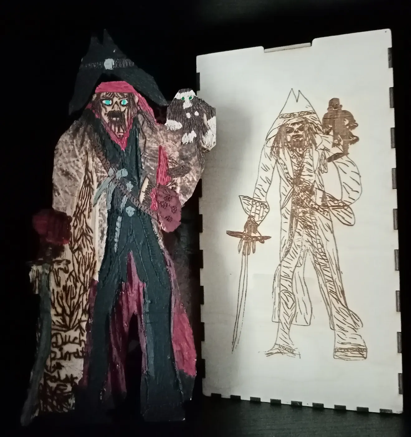 Pomalowana figurka pirata z małpką na ramieniu stoi obok jasnej drewnianej skrzynki; na wieku skrzynki jest ten sam pirat narysowany kreską