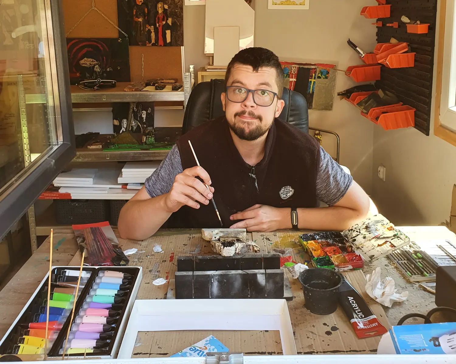 Michał Walczyk maluje przy biurku w pracowni, przed nim pastele i farby