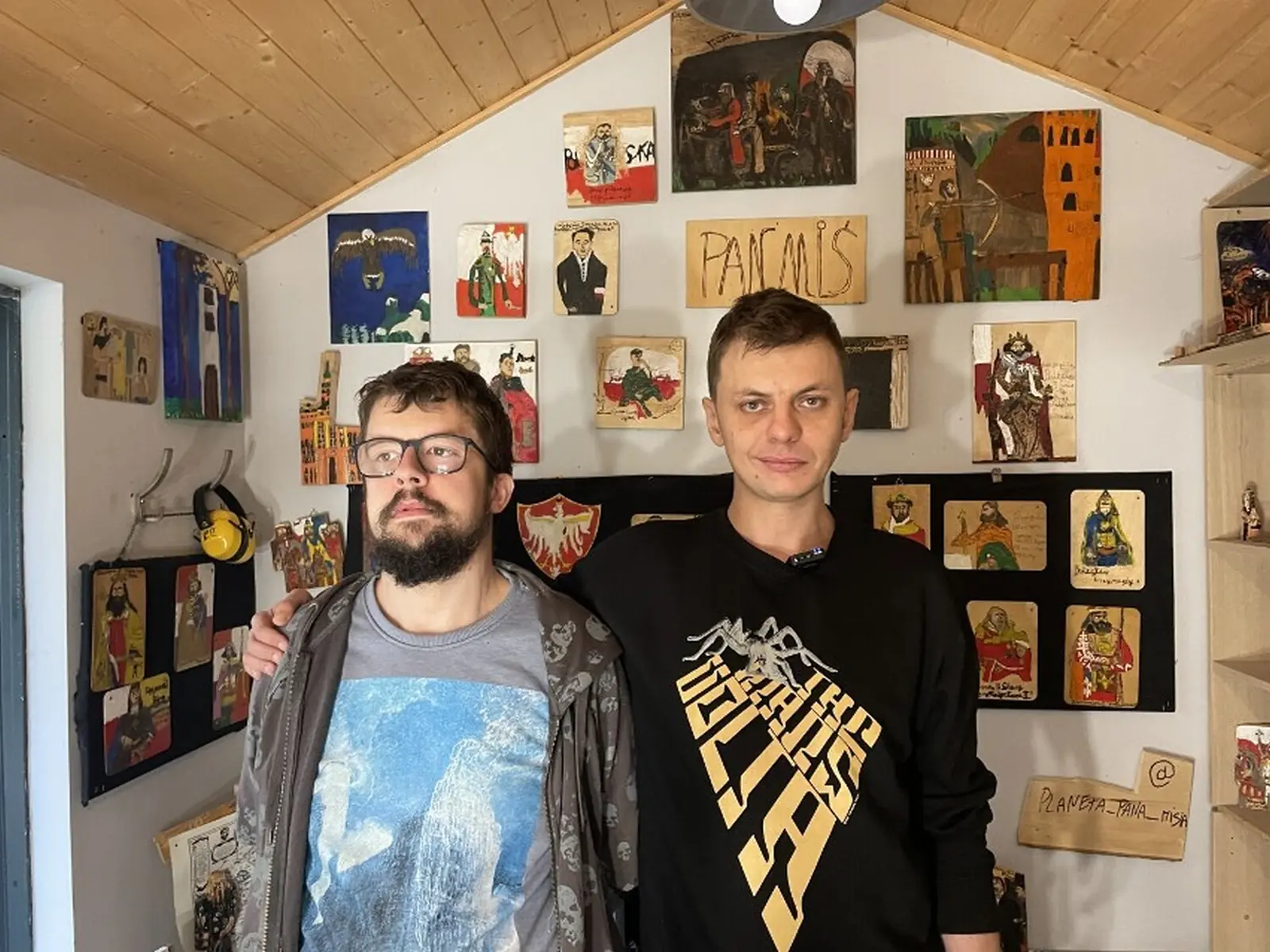 Dwóch mężczyzn stoi w pracowni na tle ściany z obrazami; z lewej Michał Walczyk